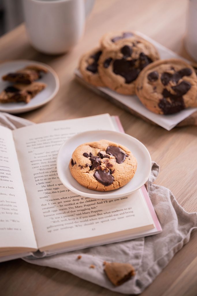 Warm chocolate chip cookie served on a small plate resting over an open book, with a tray of cookies and a coffee cup in the background. Photographed by Lou Carruthers, food and hospitality photographer.