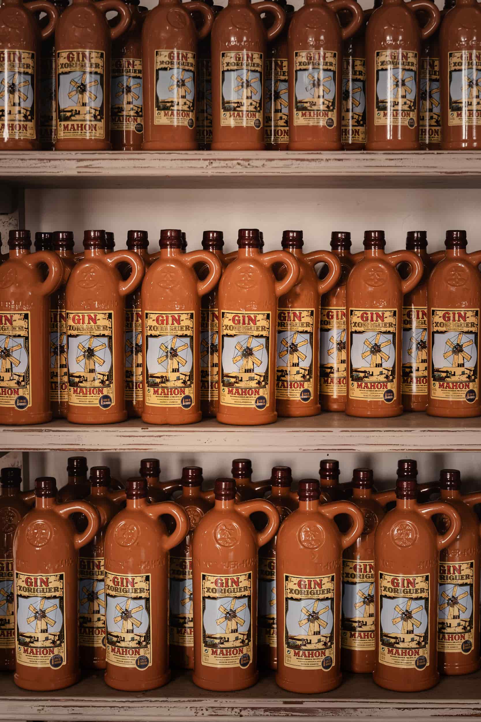 Rows of terracotta bottles of Xoriguer gin neatly arranged on shelves at Destilería Xoriguer, Menorca, showcasing the island’s local spirit. Photographed by Lou Carruthers, hospitality and food photographer.
