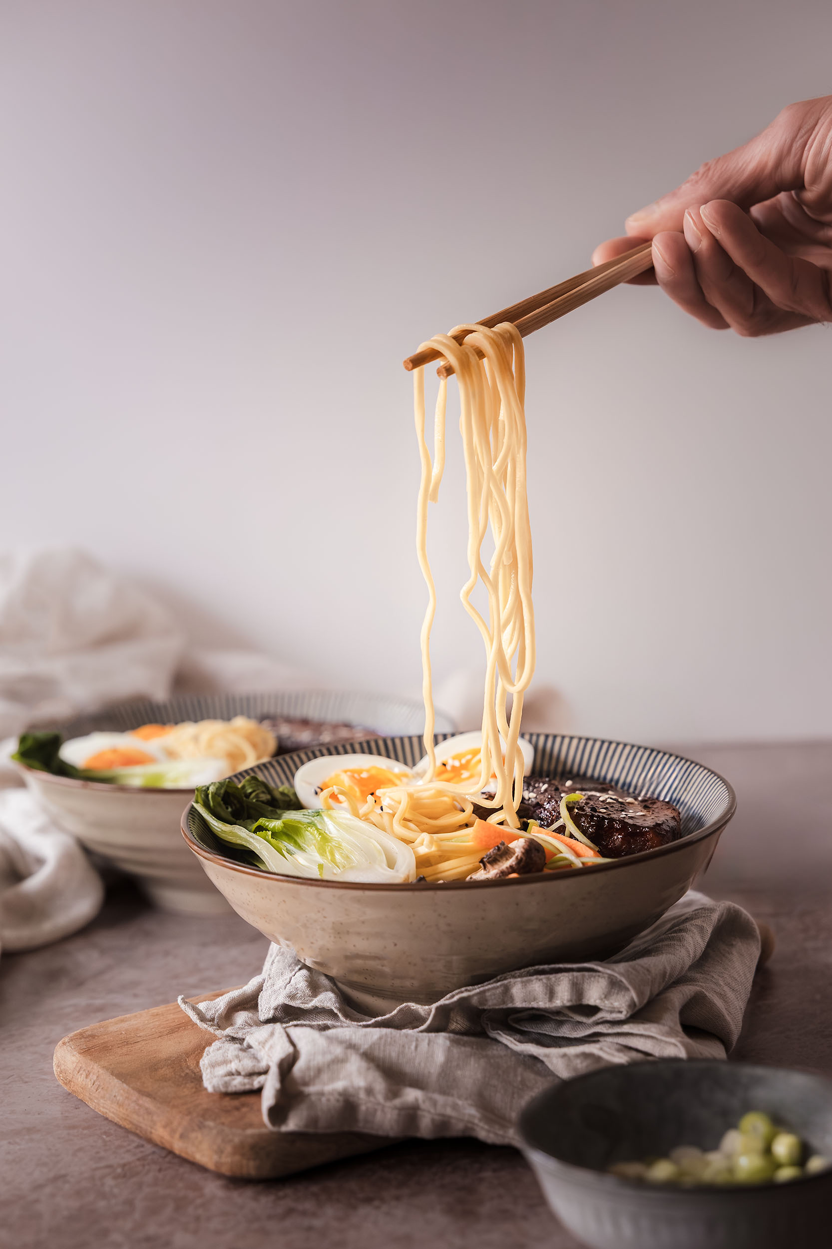 Hand lifting noodles with chopsticks from a steaming ramen bowl, styled with Asian cuisine ingredients including soft-boiled eggs, pak choi, mushrooms and sesame seeds. Captured as part of restaurant food photography by Lou Carruthers, hospitality and food photographer.