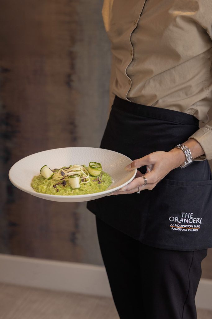 Lou Carruthers – Server at The Orangery, Siddington Park, presenting a vibrant green pea and courgette risotto garnished with micro herbs, captured in a clean hospitality service setting.