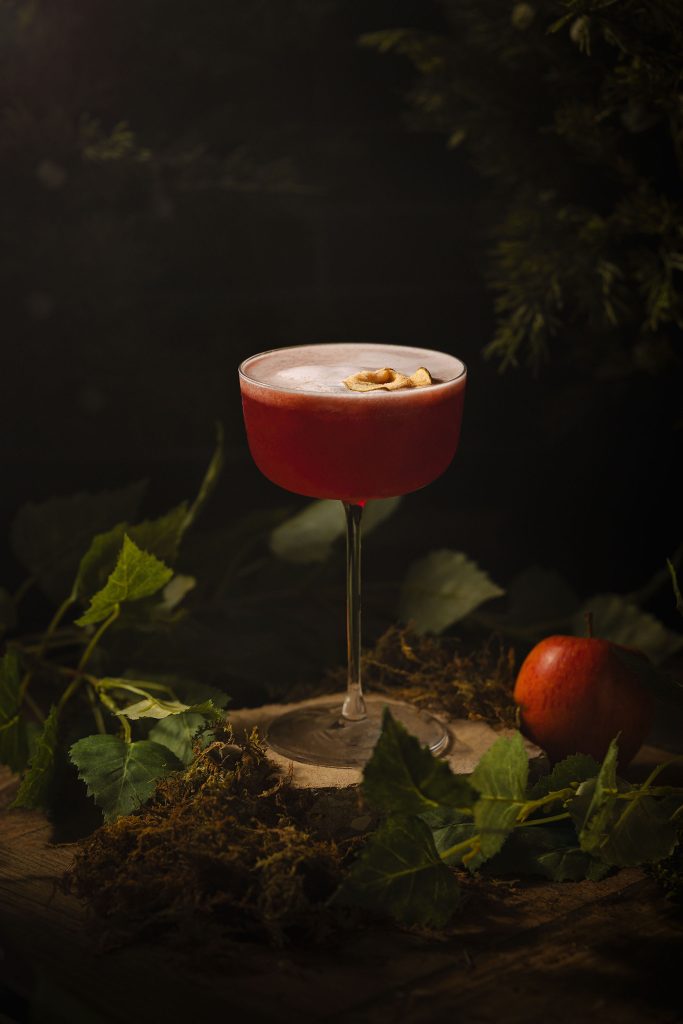 Evil Queen cocktail photographed by Cotswold Food and Drink Photographer, Lou Carruthers. A deep ruby-red drink in a coupe glass surrounded by moss and forest leaves, softly lit to evoke an enchanted woodland. Inspired by the fairytale’s poisoned apple and crafted to capture dark beauty and temptation.