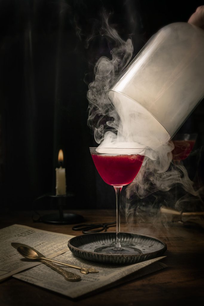 Smoke-filled cocktail photograph featuring a deep red drink revealed beneath a glass cloche, styled with antique gold cutlery and candlelight for a cinematic, moody atmosphere. Photographed by Lou Carruthers, food and drink photographer.