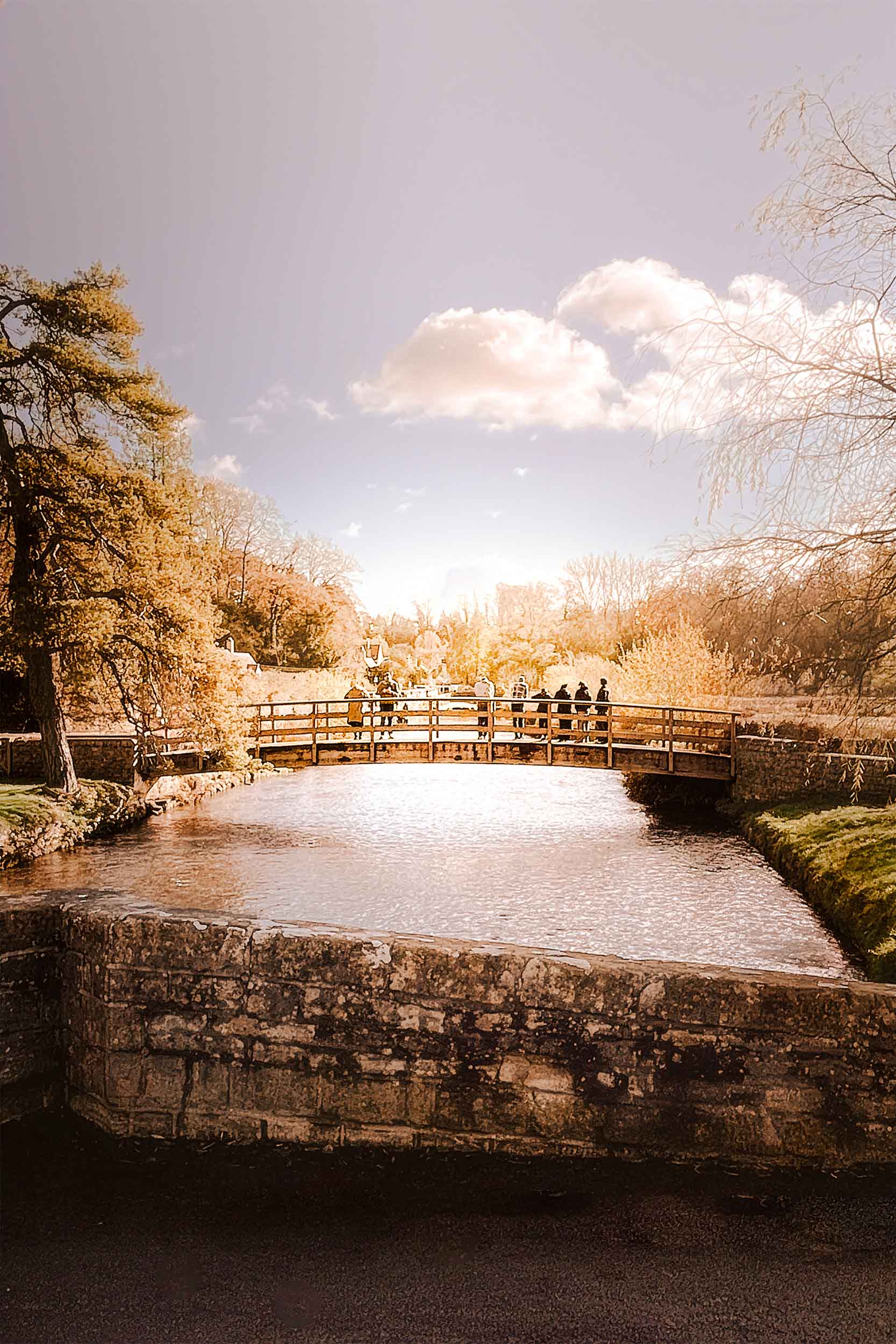 Visitors crossing the wooden bridge over the River Coln in Bibury during golden hour, captured by Lou Carruthers for the Taste • Light • Story Spring Retreat in the Cotswolds.