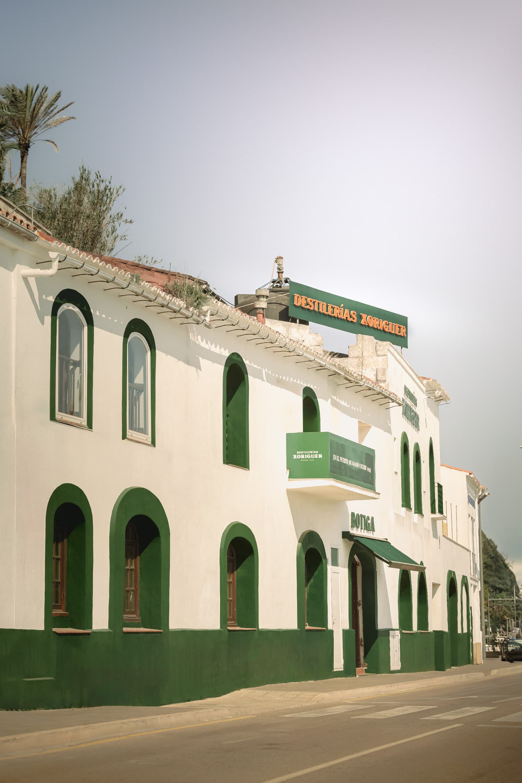 The whitewashed façade of the Xoriguer gin distillery in Mahón, Menorca, with arched green windows and the iconic sign above the entrance. Photographed by Lou Carruthers, food and hospitality photographer.