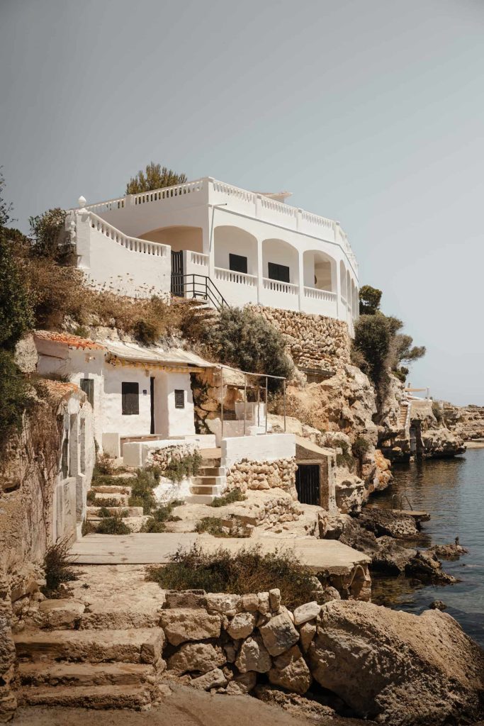 Whitewashed coastal building perched above the rocky shoreline at Cala Alcaufar, Menorca, overlooking the clear Mediterranean waters. Photographed by Lou Carruthers, hospitality and travel photographer.