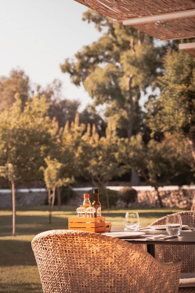 Outdoor dining table set for breakfast under dappled sunlight at Malbuger Nou in Menorca, with olive oil, vinegar, and condiments in a wooden crate, surrounded by wicker chairs and lush garden views. Photographed by Lou Carruthers, specialist in food and hospitality photography.