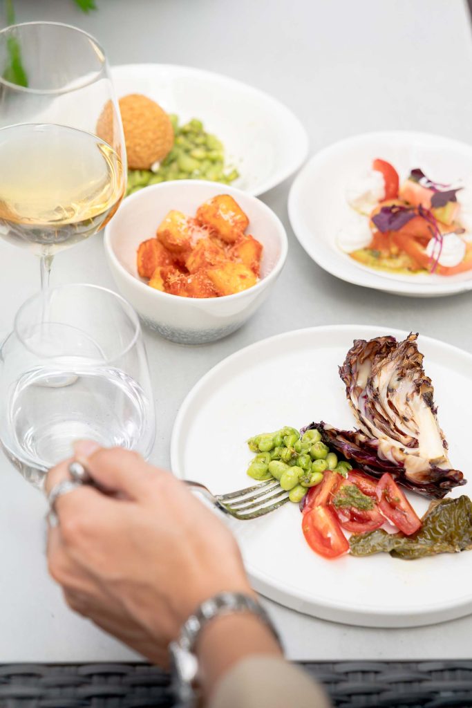 Outdoor dining scene with small plates of Mediterranean dishes, including grilled vegetables, roasted potatoes, and salads, paired with white wine and water. Photographed by Lou Carruthers, hospitality and food photographer.
