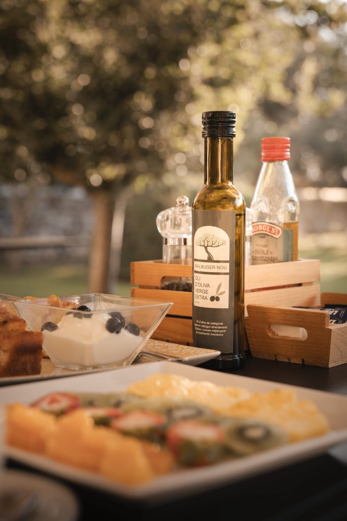 Close-up of a rustic outdoor breakfast table featuring a bottle of Malbuger Nou extra virgin olive oil, natural yogurt with blueberries, and fresh fruit in soft morning light — styled for a relaxed Mediterranean setting in Menorca. Captured by Lou Carruthers, hospitality and food photographer.