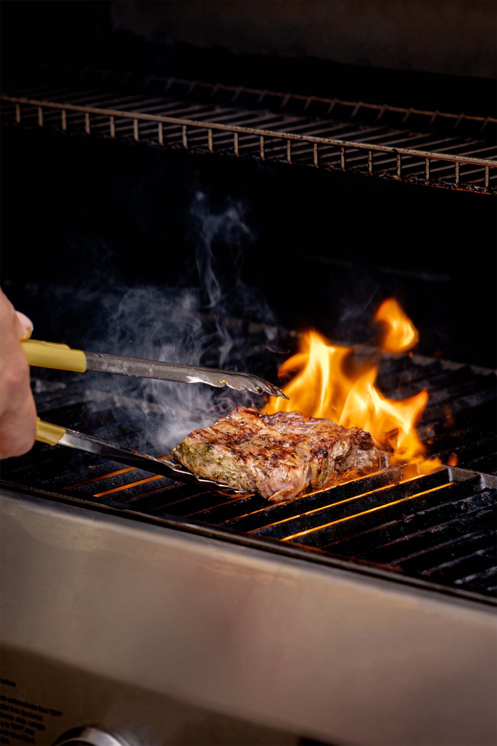 Chef grilling seasoned meat over open flames on a barbecue grill, capturing the heat and smoke of outdoor cooking for a hospitality menu. Photographed by Lou Carruthers, hospitality and food photographer.
