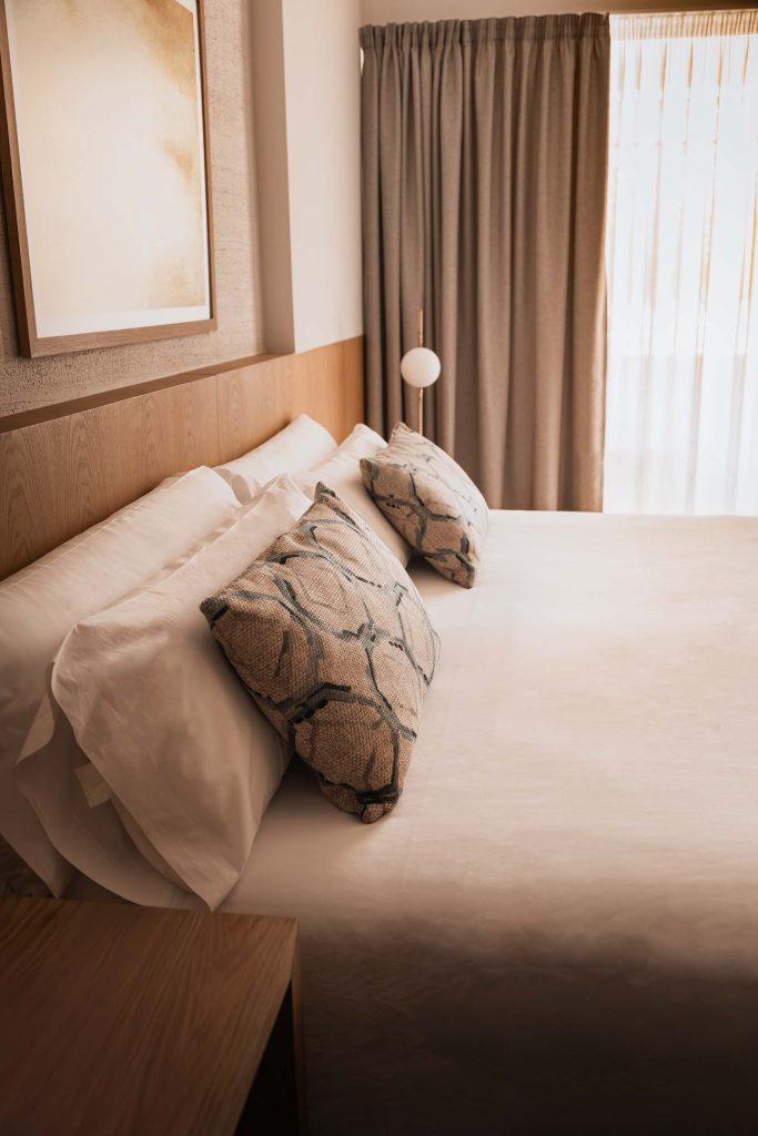 Minimalist hotel bedroom at Malbuger Nou in Menorca with crisp white linens, textured patterned cushions, and warm natural light filtering through soft curtains. Photographed by Lou Carruthers, hospitality and travel photographer.