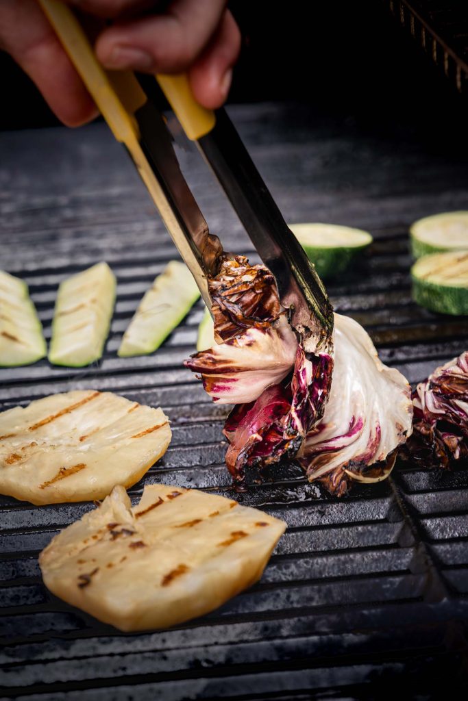 Close-up of grilled vegetables including celeriac slices and radicchio on a barbecue grill, prepared for a summer hospitality menu. Photographed by Lou Carruthers, hospitality and food photographer.