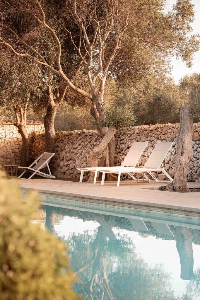 Tranquil poolside scene at Malbuger Nou in Menorca with white sun loungers beneath olive trees, surrounded by a traditional stone wall in soft golden light. Photographed by Lou Carruthers, hospitality and travel photographer.
