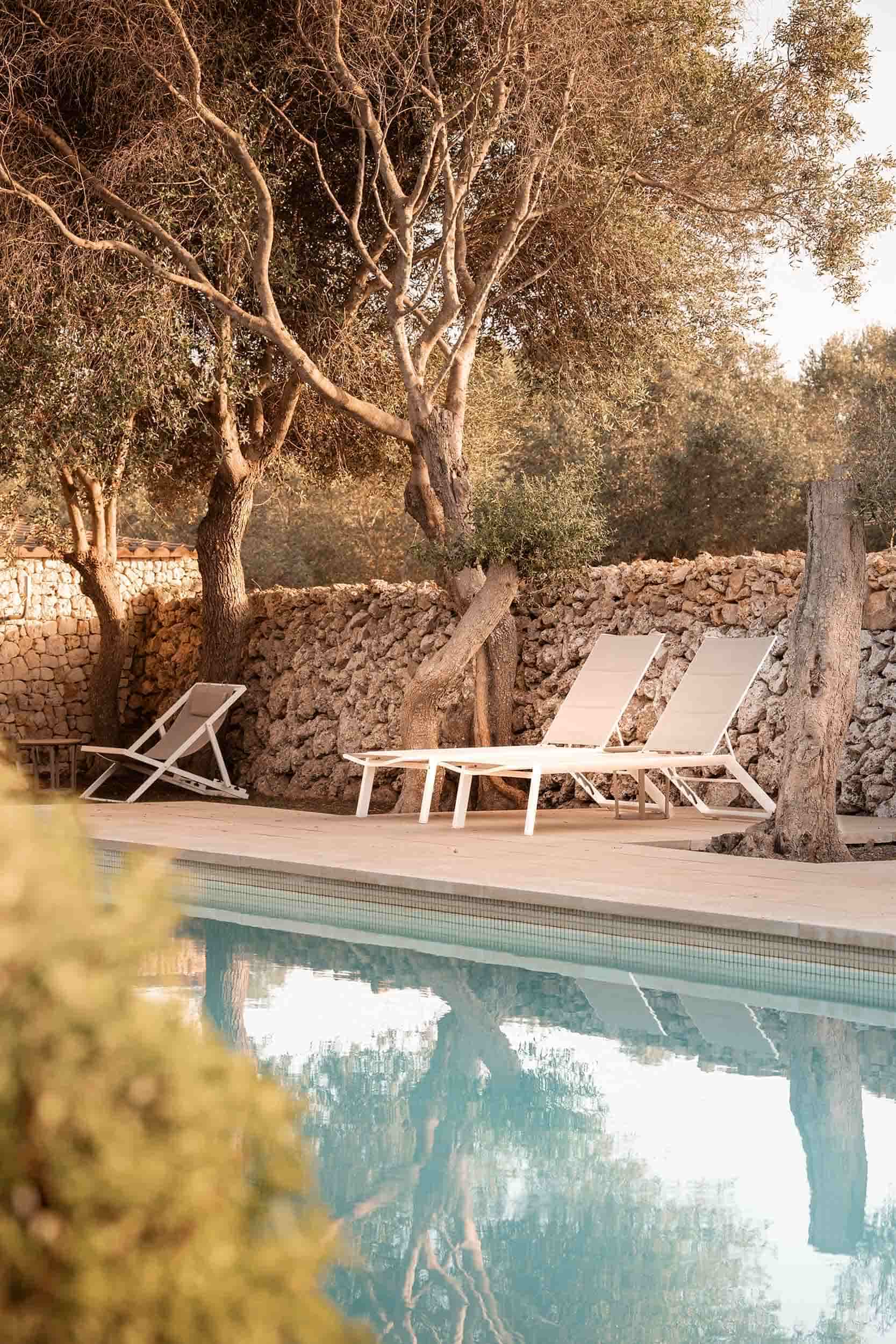 Tranquil poolside scene at Malbuger Nou in Menorca with white sun loungers beneath olive trees, surrounded by a traditional stone wall in soft golden light. Photographed by Lou Carruthers, hospitality and travel photographer.