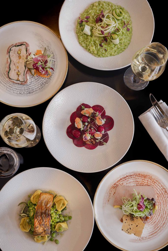 Overhead view of a gourmet tasting menu featuring colourful seasonal dishes, including beetroot salad, seafood terrine, and pasta with fresh herbs, in a fine dining setting. Photographed by Lou Carruthers, hospitality and food photographer.