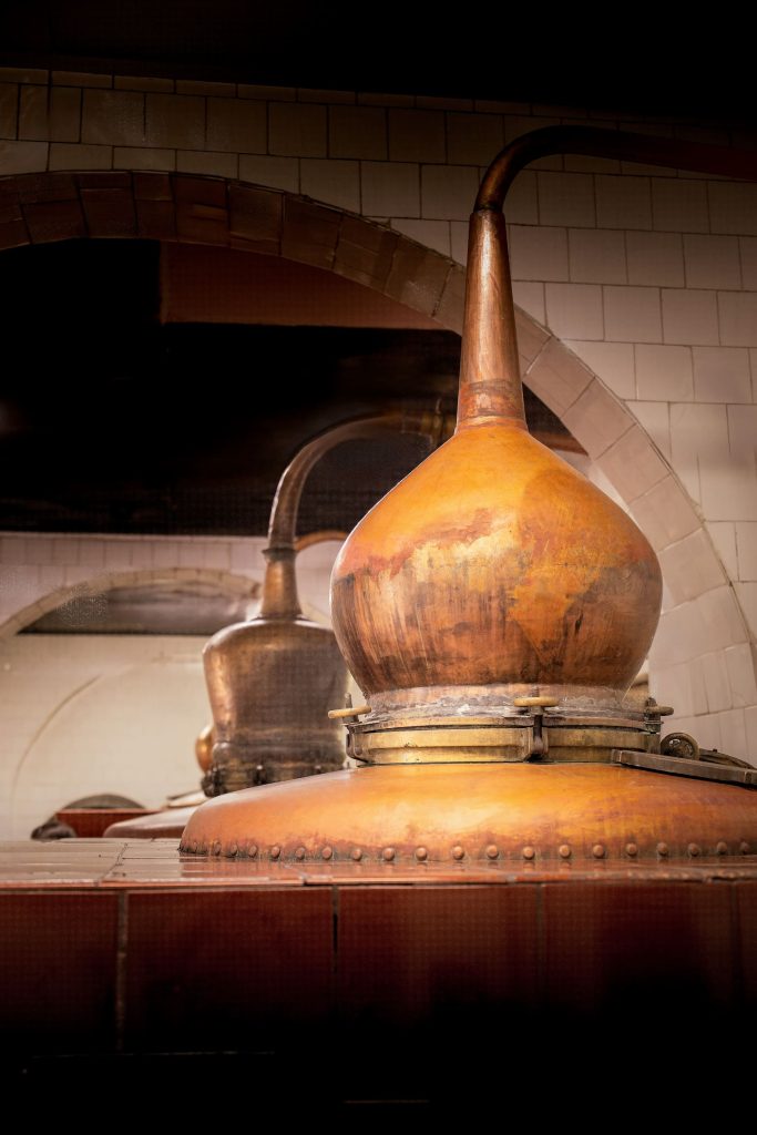 A close view of the burnished copper still at Xoriguer distillery, framed by white tiles and centuries of tradition. Photography by Lou Carruthers. Photographed by Lou Carruthers, food and hospitality photographer.