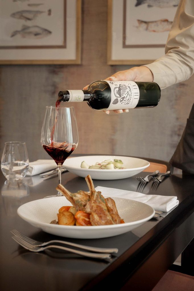 Sommelier pouring red wine into a glass beside a roasted lamb rack with seasonal vegetables in an upscale restaurant. Captured by Lou Carruthers, specialist in hospitality and food photography.