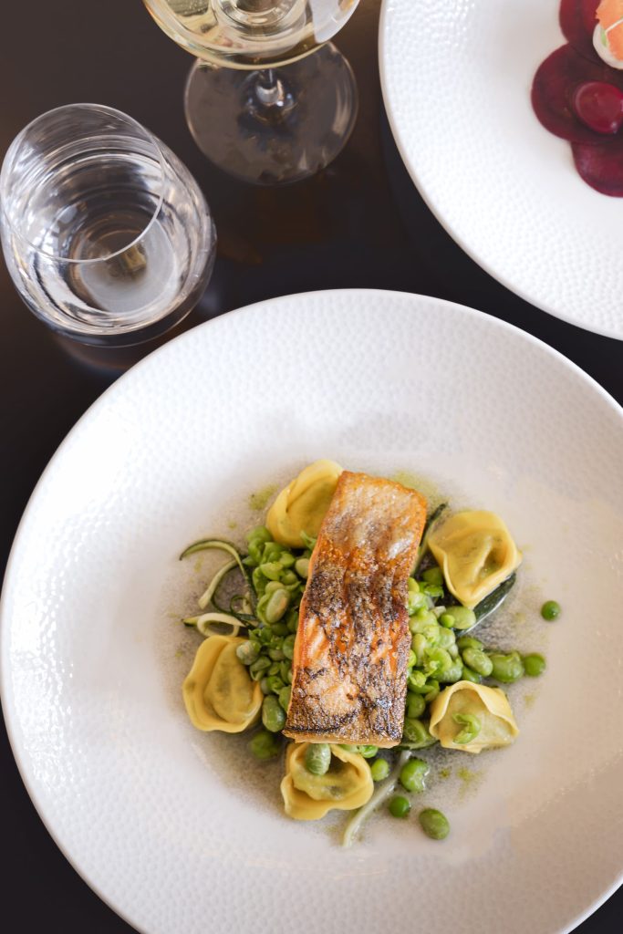 Overhead shot of pan-seared salmon served with tortellini, peas and seasonal greens at The Orangery, Siddington Park, with wine glasses styled on the table