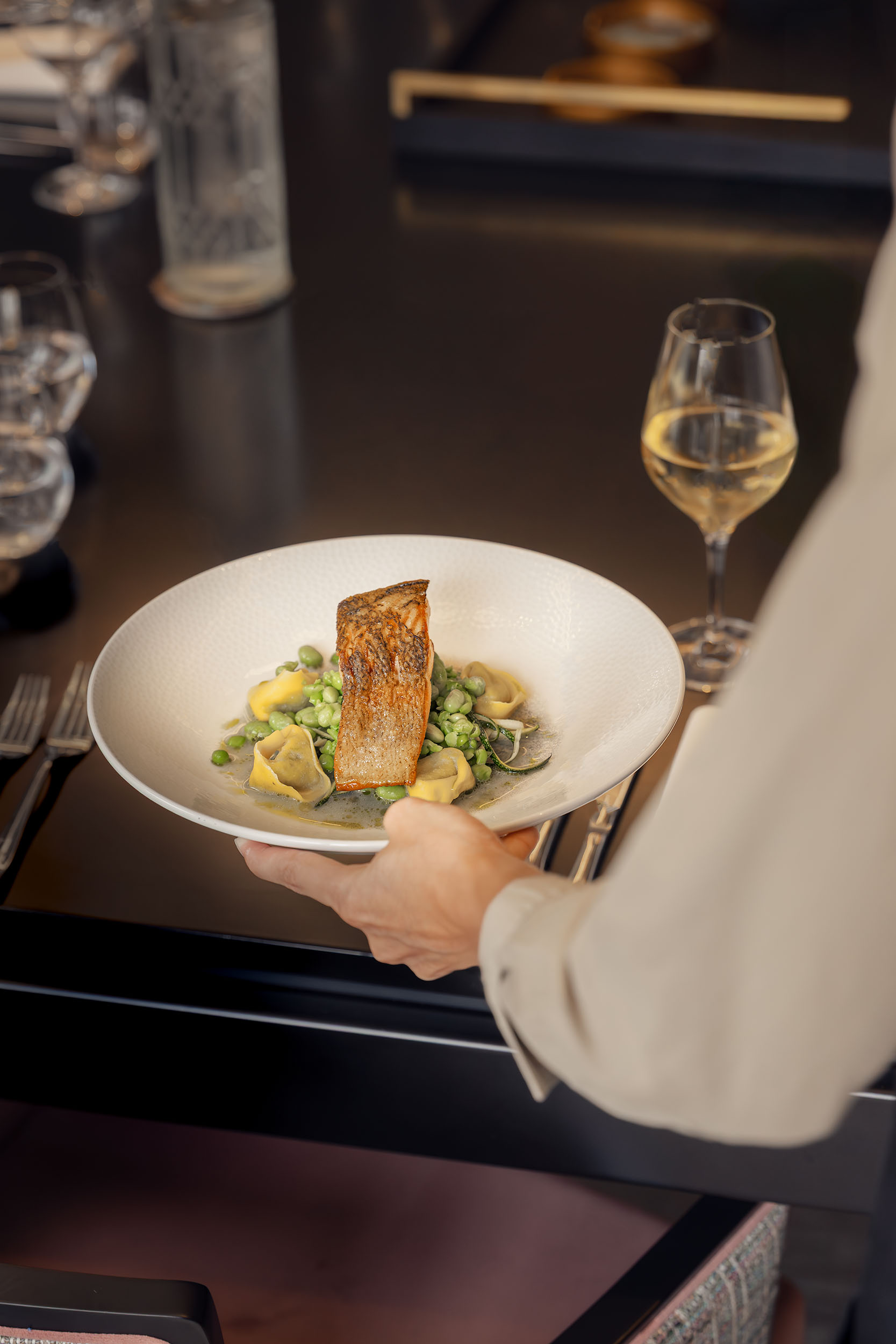 A chef serves a seared salmon fillet with seasonal greens and a glass of white wine at a fine dining restaurant table, styled for an elegant hospitality setting. Photographed by Lou Carruthers, hospitality and food photographer.