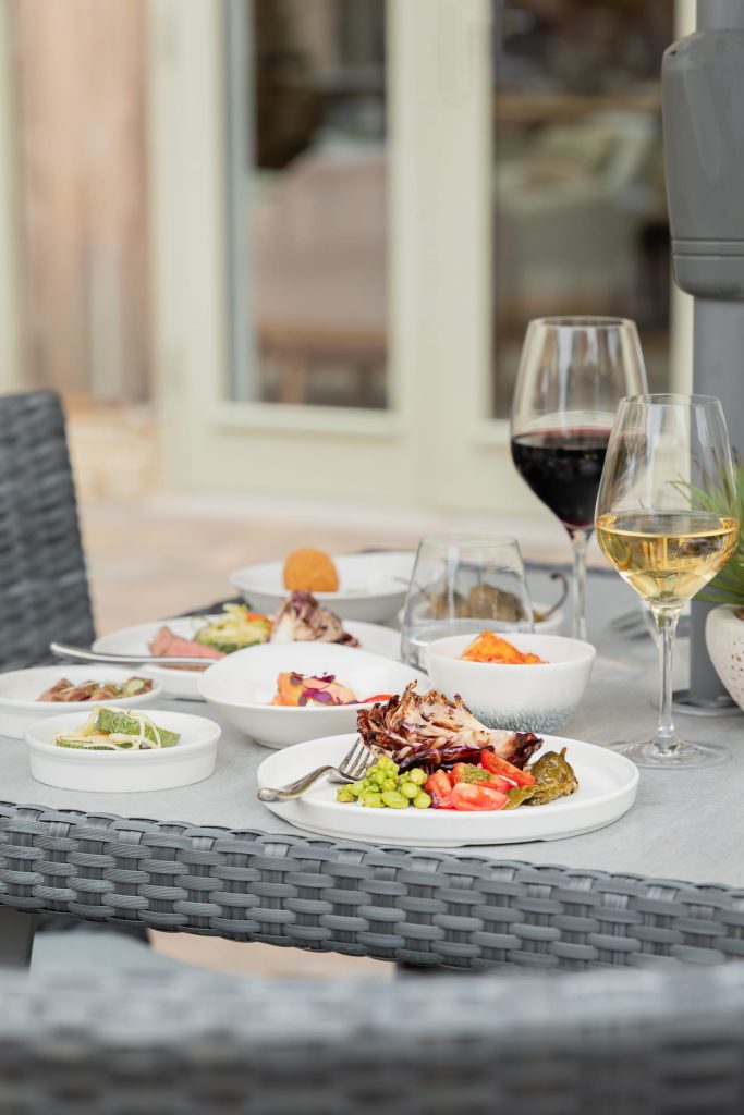 ummer dining setup at Siddington Park, featuring shared small plates, fresh seasonal dishes and glasses of white and red wine on the terrace outside The Orangery.