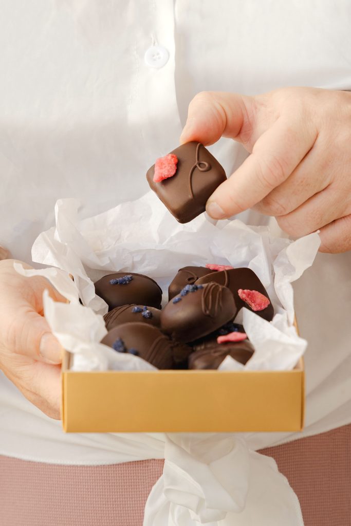 Close-up of a person holding an open box of luxury artisan dark chocolates, each decorated with crystallised petals, with one chocolate being lifted from the box. Photographed by Lou Carruthers, food and hospitality photographer.