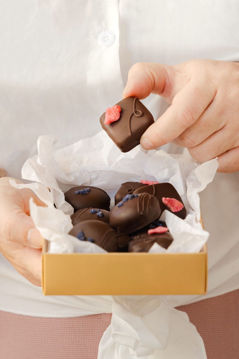 Close-up of a person holding an open box of luxury artisan dark chocolates, each decorated with crystallised petals, with one chocolate being lifted from the box. Photographed by Lou Carruthers, food and hospitality photographer.