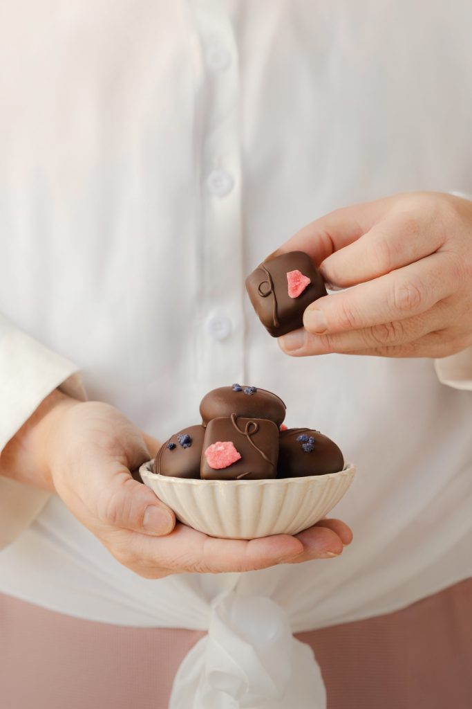 Hand holding a rose cream chocolate from a ceramic bowl filled with artisan chocolates decorated with crystallised petals. Photographed by Lou Carruthers, food and hospitality photographer.