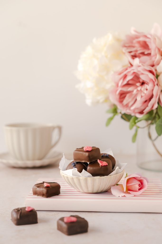 Luxury artisan dark chocolates topped with crystallised rose petals, styled in a delicate ceramic bowl beside a pink rose and teacup, creating an elegant afternoon tea scene. Photographed by Lou Carruthers, food and hospitality photographer.