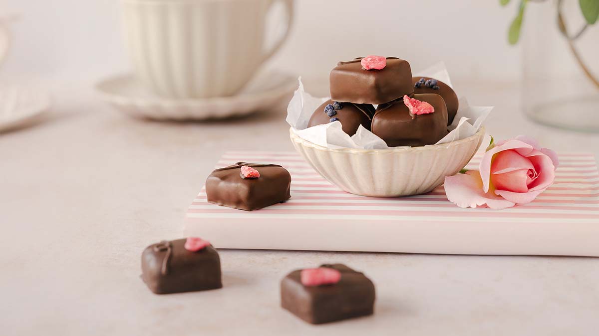 Luxury artisan dark chocolates topped with crystallised rose petals, styled in a delicate ceramic bowl beside a pink rose and teacup, creating an elegant afternoon tea scene. Photographed by Lou Carruthers, food and hospitality photographer.
