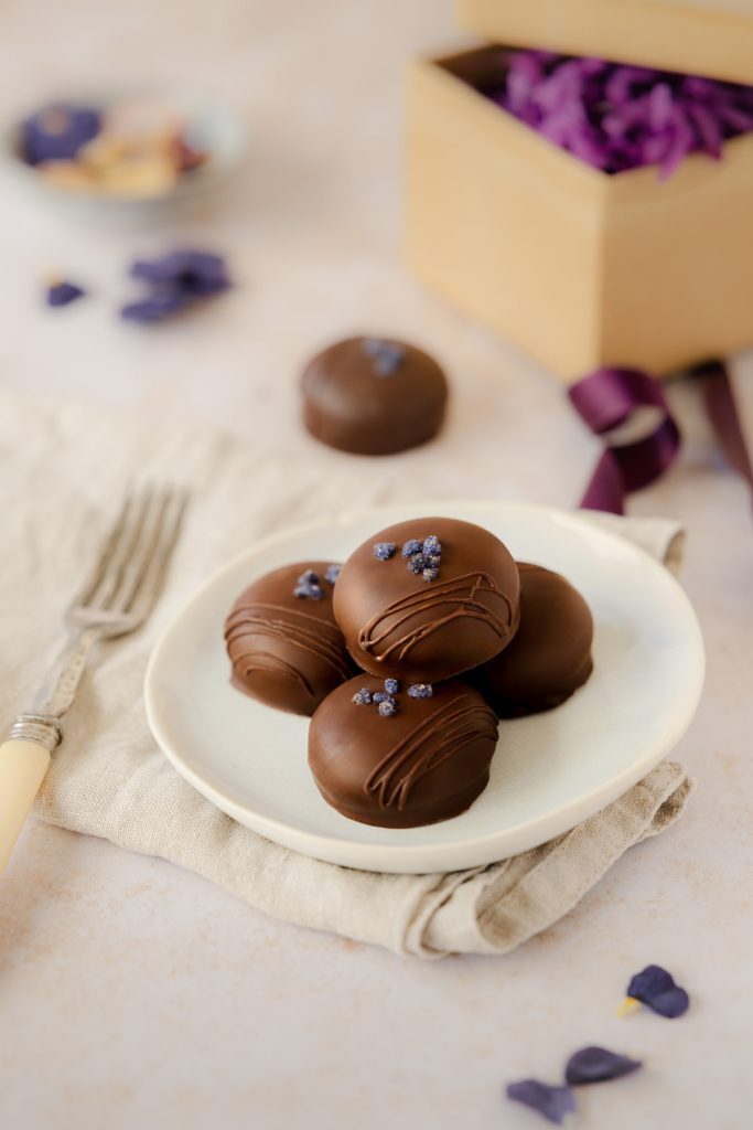 Plate of artisan violet cream chocolates, decorated with crystallised petals and styled with a ribbon and gift box in the background. Photographed by Lou Carruthers, food and hospitality photographer.