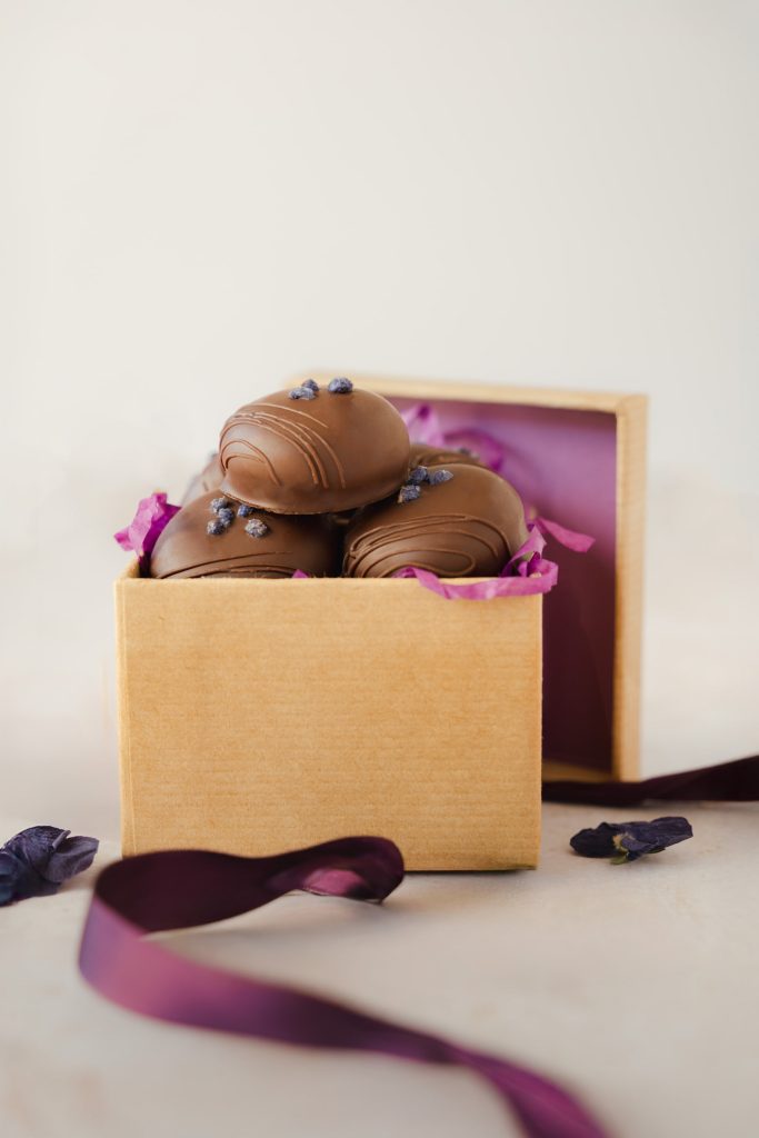 Gift box of artisan violet cream chocolates with crystallised decorations, styled with purple ribbon and tissue paper. Photographed by Lou Carruthers, food and hospitality photographer.