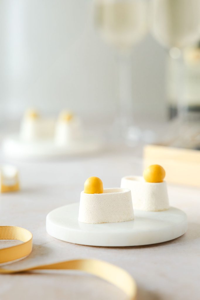 Elegant lemon meringue petit fours with glossy yellow spheres on top of white mousse bases, styled on marble plates with champagne glasses in the background. Photographed by Lou Carruthers, food and hospitality photographer.