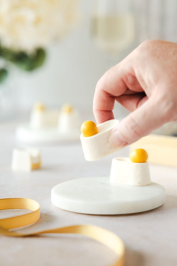 Hand serving a lemon meringue petit four, featuring a bright yellow sphere on a white mousse base, luxury artisan desserts styled for gifting. Photographed by Lou Carruthers, food and hospitality photographer.