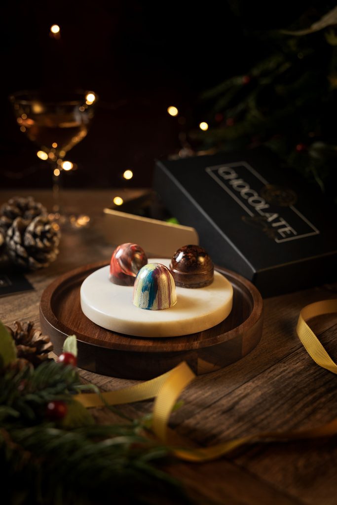 Artisan chocolate bonbons from Chocolate Bliss displayed on a marble plate with festive décor and warm golden bokeh, styled and photographed by Lou Carruthers.
