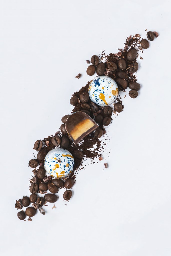 Luxury dark chocolate rose creams decorated with crystallised petals, styled with dried pink and purple flower petals on a white background. Photographed by Lou Carruthers, artisan food photographer.