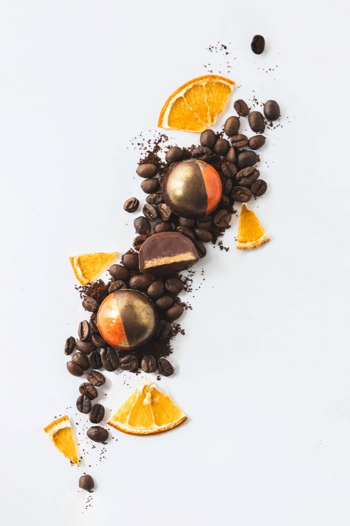 Luxury artisan chocolate bonbons in metallic orange and gold, styled with coffee beans and dried orange slices on a white background. Photographed by Lou Carruthers, food and hospitality photographer.