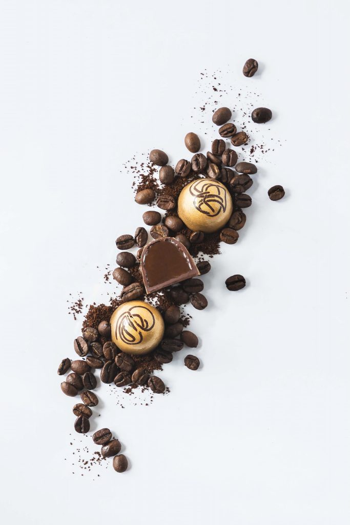 Golden artisan coffee bonbons styled with roasted coffee beans and cocoa powder on a white background. Photographed by Lou Carruthers, food and hospitality photographer.