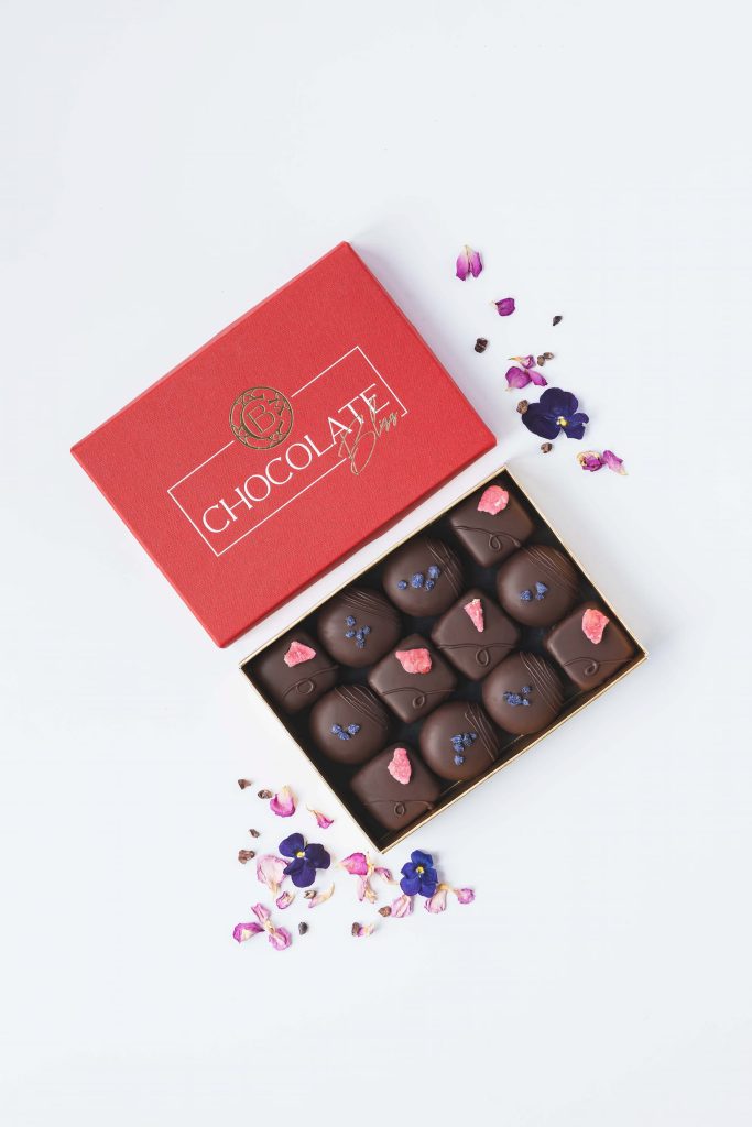 Luxury presentation box of artisan rose and violet cream chocolates, decorated with crystallised petals and styled with scattered flower petals. Photographed by Lou Carruthers, food and hospitality photographer.