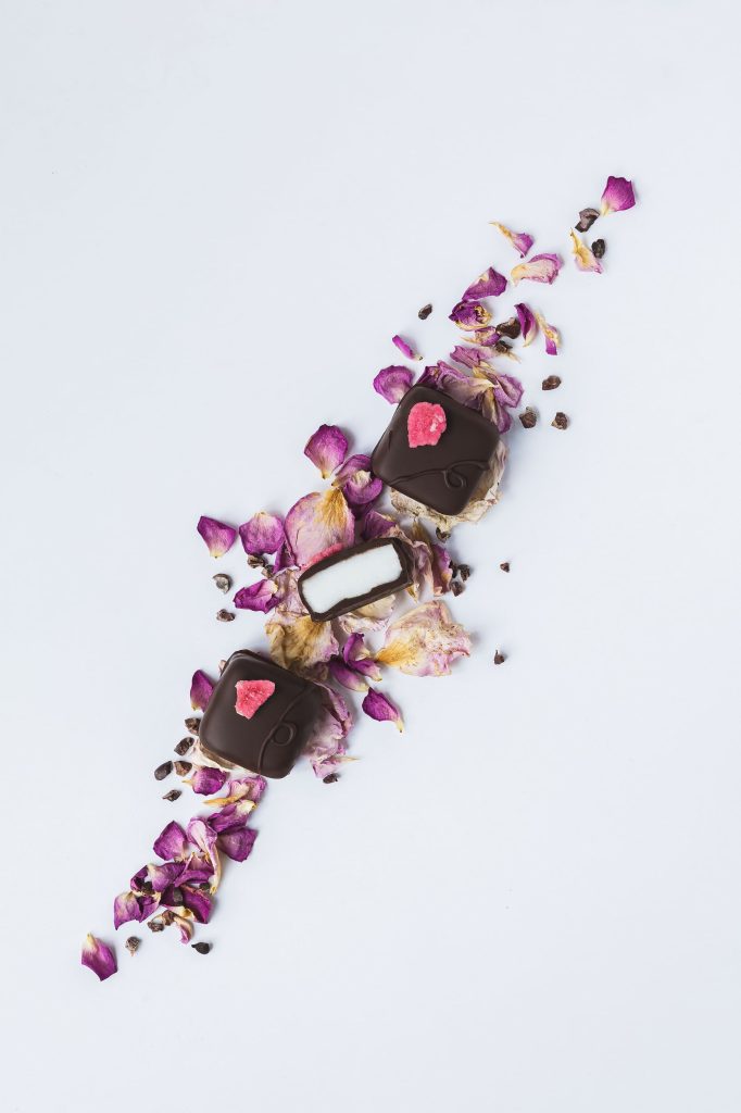 Artisan dark chocolate rose creams with crystallised petal decoration, styled on dried rose petals with one chocolate cut open to show the creamy centre. Photographed by Lou Carruthers, food and hospitality photographer.