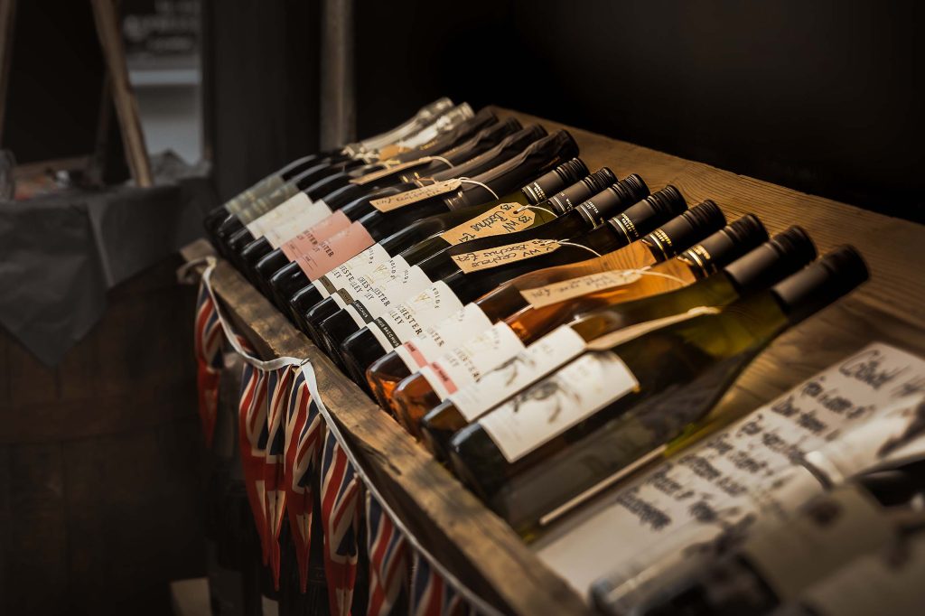 Row of craft wine bottles with handwritten labels displayed on a striped cloth-covered wooden table, captured in warm natural light with soft shadows - artisan drinks product photography by Lou Carruthers