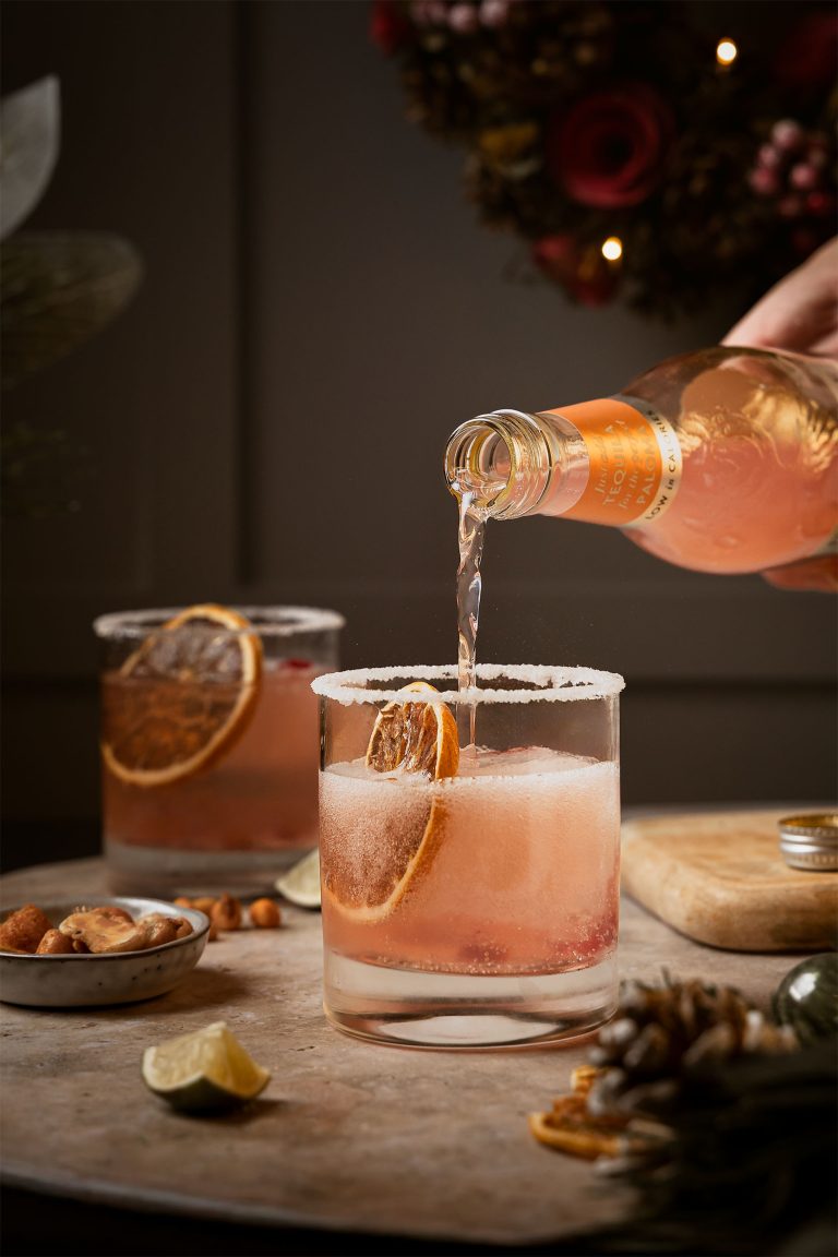 Hand pouring Fever-Tree Pink Grapefruit Soda into cocktail glass with salted rim and fresh grapefruit garnish for festive Paloma drink, photographed by Lou Carruthers, drinks photographer