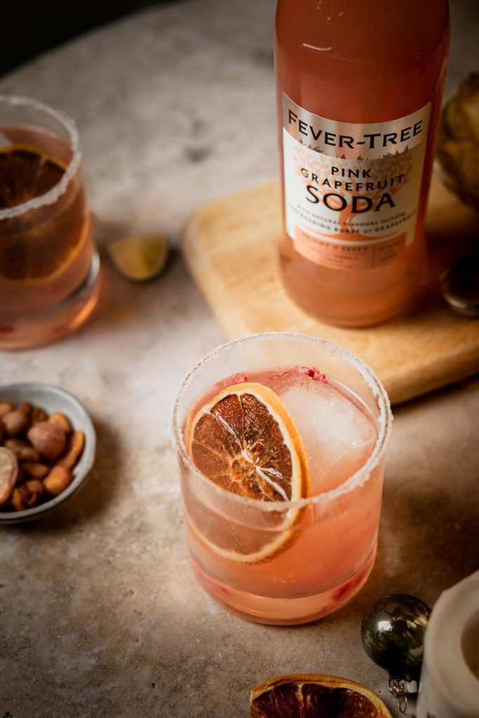 Two festive Paloma cocktails with dried orange slices and a Fever-Tree Pink Grapefruit Soda bottle styled among winter greenery and soft candlelight by Lou Carruthers Photography