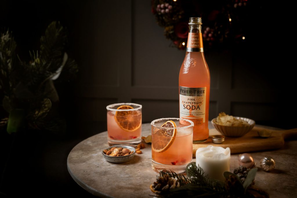 Two festive Paloma cocktails with dried orange slices and a Fever-Tree Pink Grapefruit Soda bottle styled among winter greenery and soft candlelight by Lou Carruthers Photography