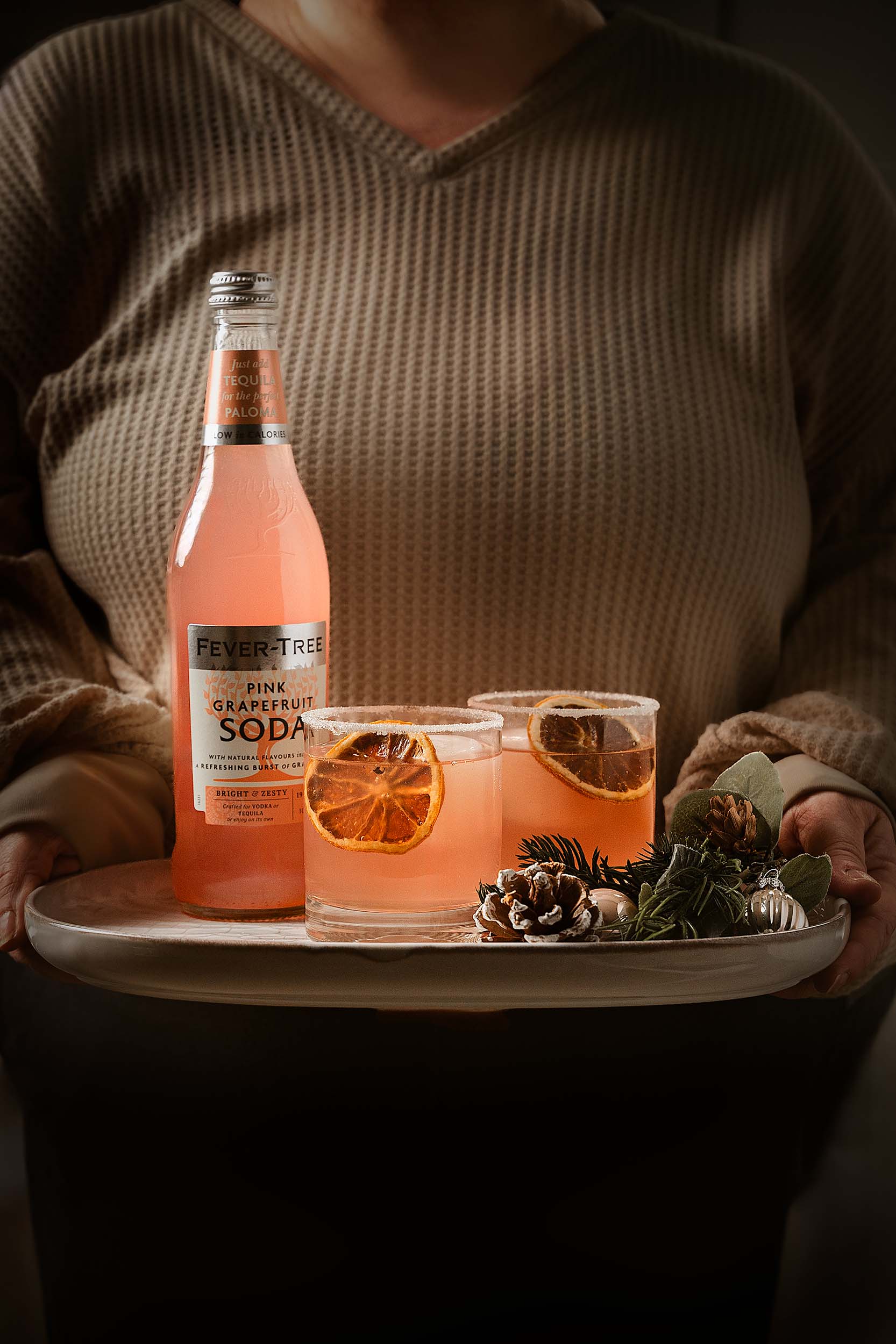 Fever-Tree Pink Grapefruit Soda bottle and two Paloma cocktails presented on a tray with pinecones and festive greenery, held by a host in warm winter light by Lou Carruthers Photography