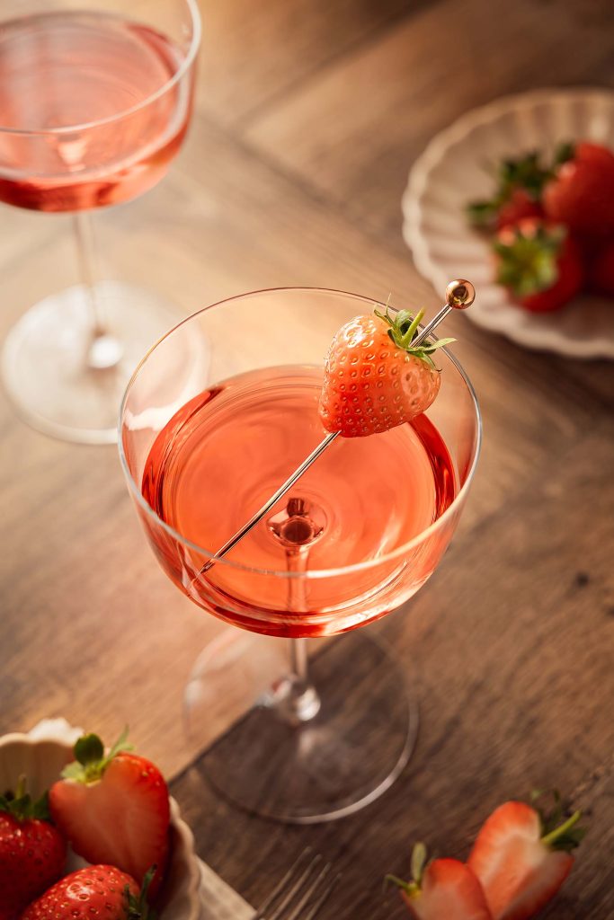 A close-up of a strawberry-topped gin cocktail in a coupe glass, surrounded by fresh sliced berries and warm afternoon light, photographed by Lou Carruthers.