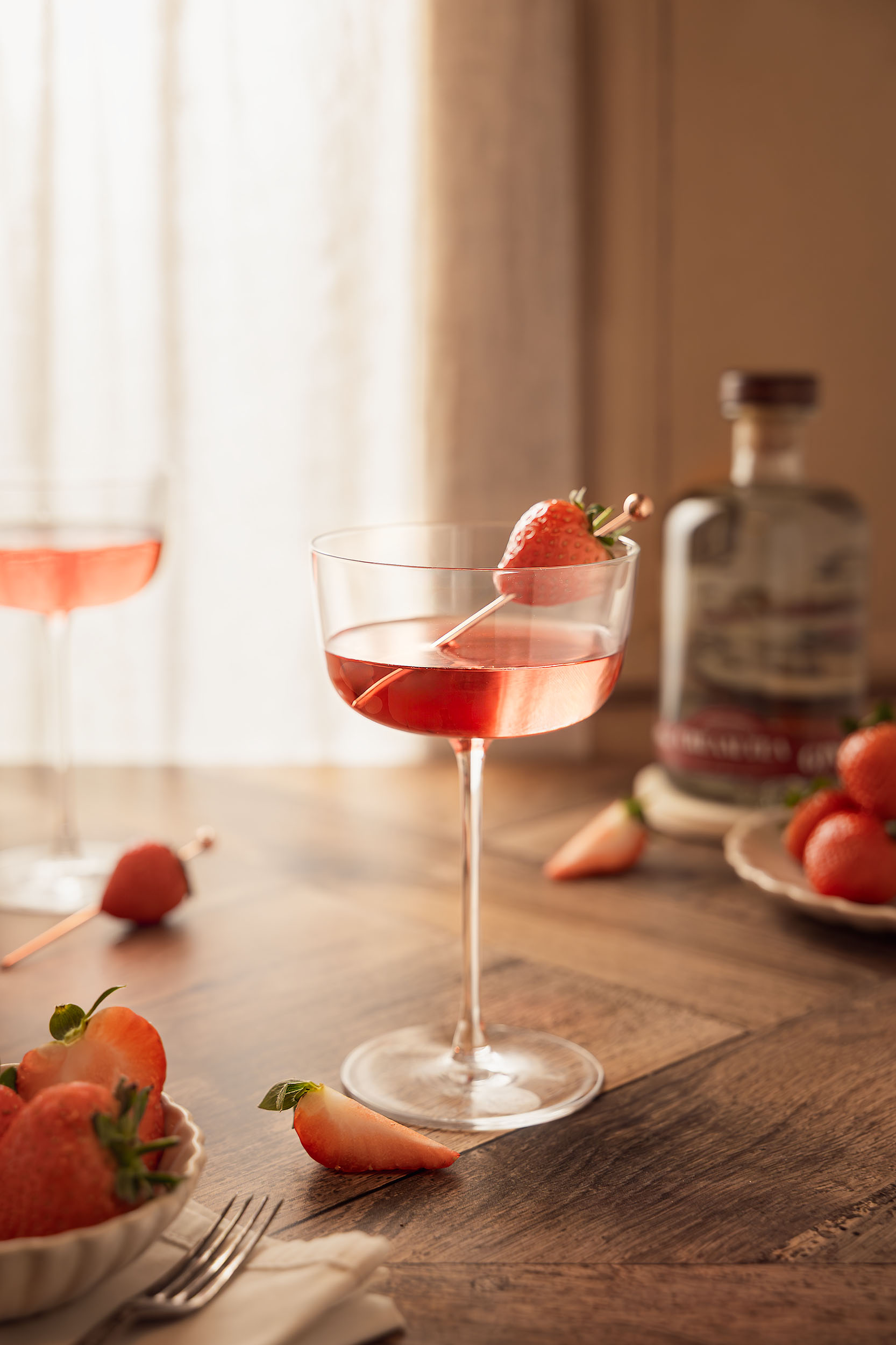A strawberry-garnished gin cocktail served in a coupe glass beside fresh berries and a Cream Tea Gin bottle, captured in soft natural light by Lou Carruthers.