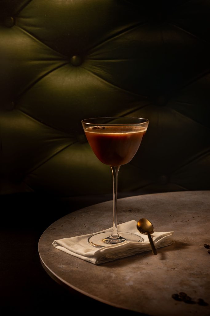 Elegant espresso martini cocktail on a marble table with soft cinematic lighting, gold spoon and folded linen napkin, styled against a dark green velvet backdrop. Photographed by Lou Carruthers, food and drink photographer for hospitality and craft brands.