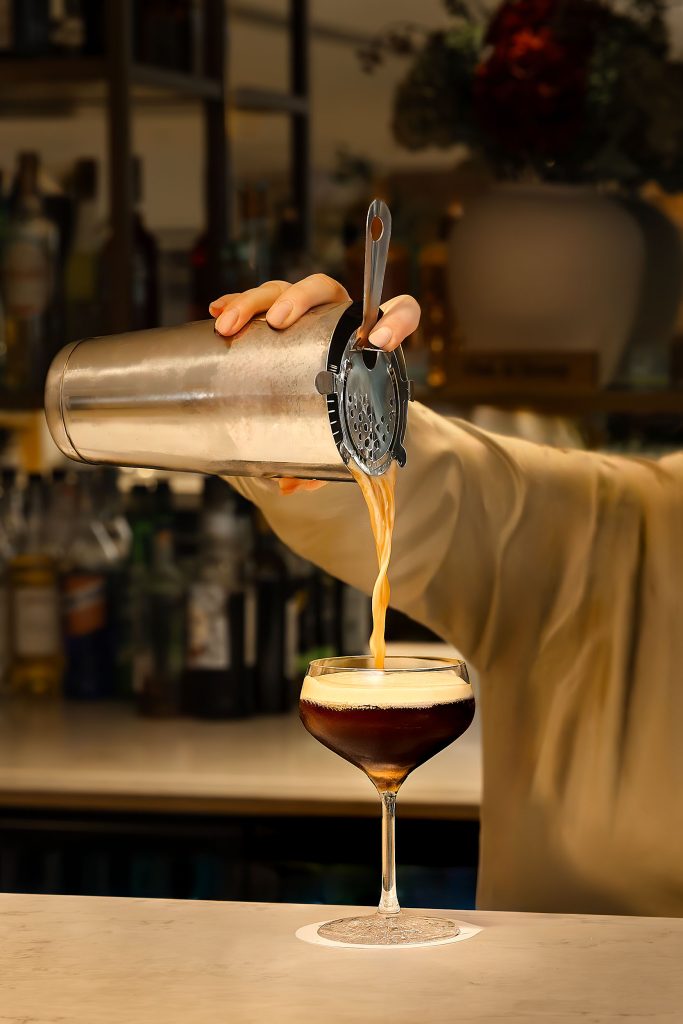 Bartender pouring a freshly shaken espresso martini into a coupe glass in a hotel bar.
