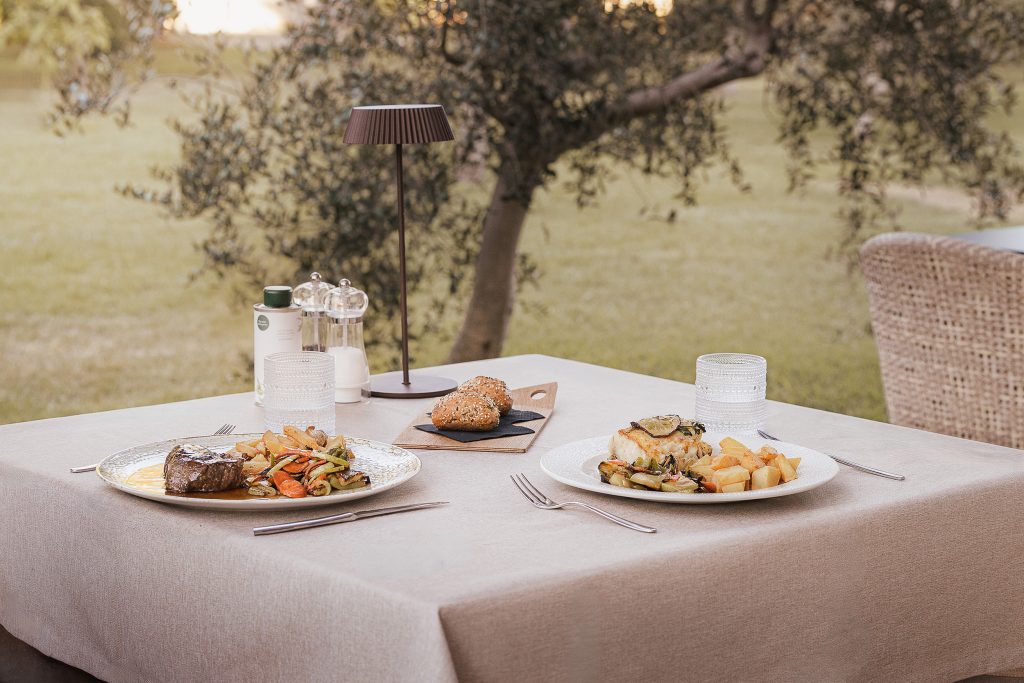 An elegant outdoor dining table at Malbuger Nou in Menorca, set with seasonal Mediterranean dishes overlooking peaceful olive trees. Soft evening light and natural surroundings create a relaxed al fresco dining experience.