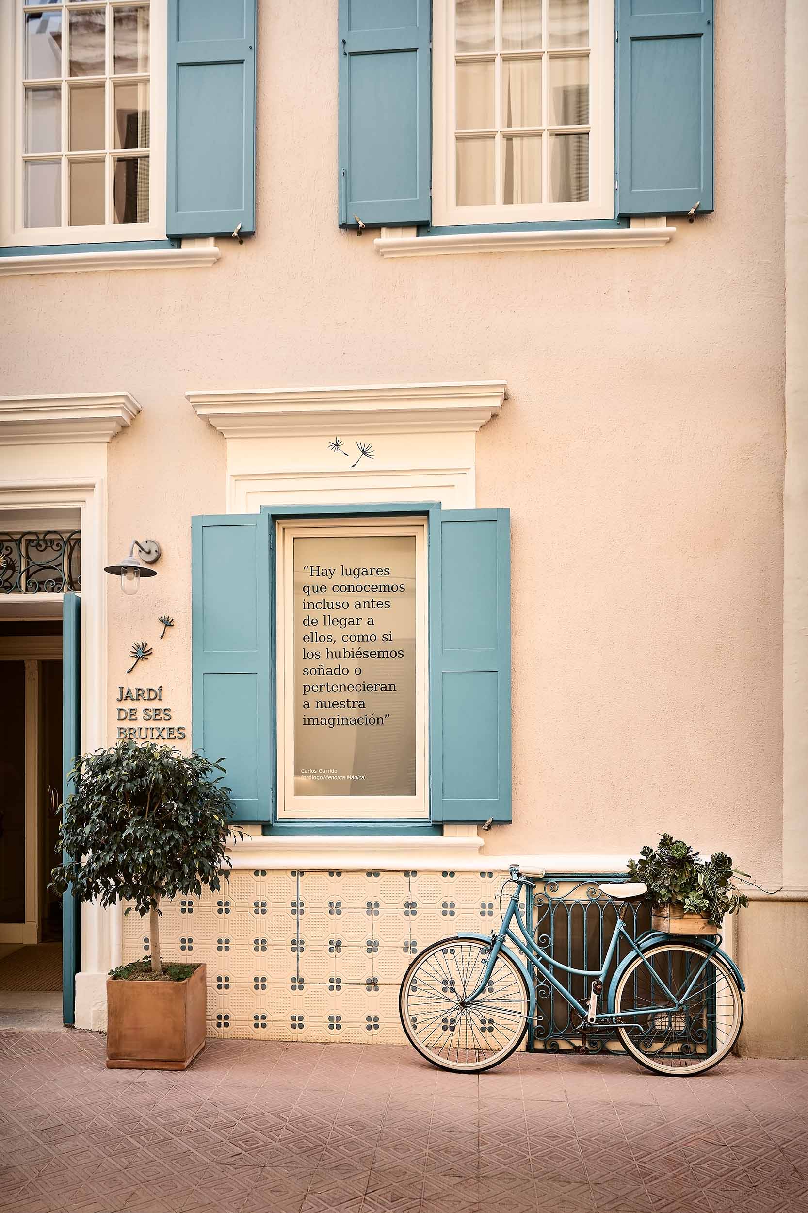 Exterior of Jardí de Ses Bruixes boutique hotel in Menorca, with blue shutters, a painted bicycle, patterned tiles and soft Mediterranean light.