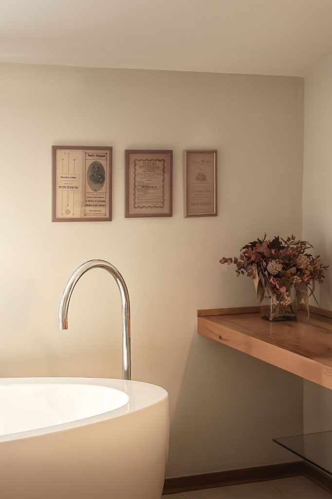 Detail of a boutique hotel bathroom in Menorca, showing a freestanding white bath, chrome tap, warm wood shelf and soft neutral walls.